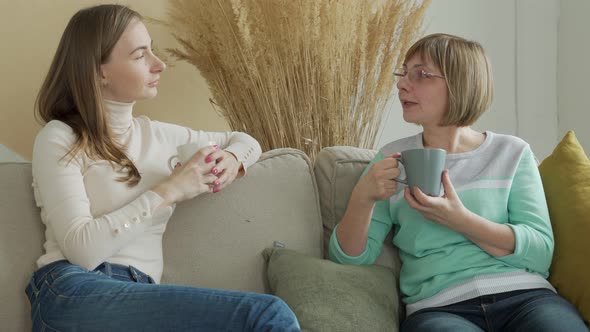 Elderly Mother with a Mug in Her Hands is Sitting on the Sofa with Her Cute Young Daughter Who is alt