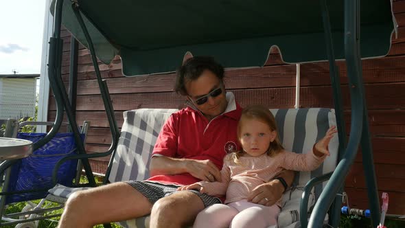 Smiling Dad With Cute Child Girl. Father And Kid Daughter Sit On Swing Set Outdoors 