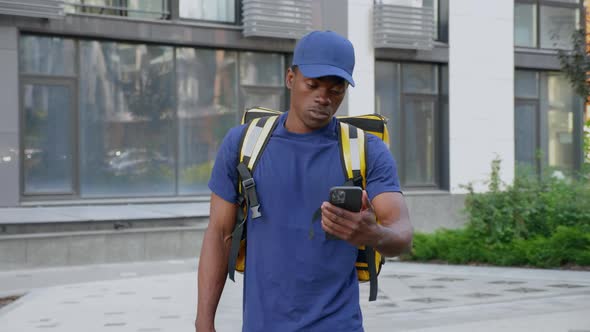 African Man Courier Delivery with Backpack Walks Street Watching in Smartphone alt