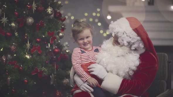 Little Girl Sitting on Santa Claus Lap in Decorated Living Room alt