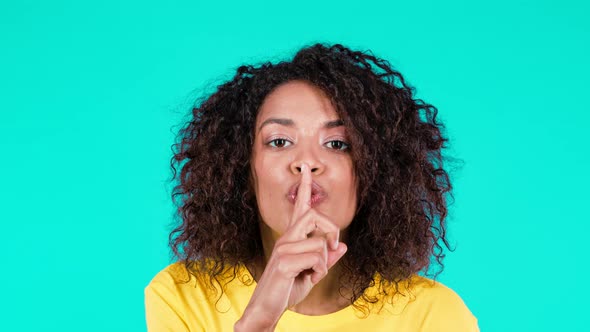 Smiling Multiethnic Woman Holding Finger on Her Lips Over Teal Background alt