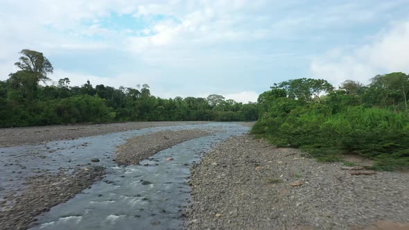 Drone shot flying over a tropical stream covered in pebbles and a lush green colored forest alt