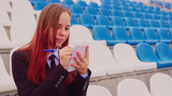 Young Woman with Notepad Pen Sitting on Stadium Bleachers Alone alt