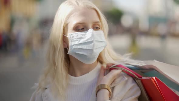 A Tired Woman in a Medical Mask Walks Down the Street with Bags After Shopping