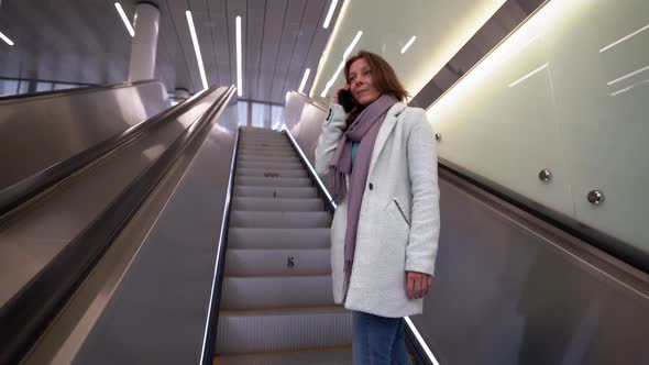 Smiling Woman Rides Up the Escalator, She Is in a Public Place, Talking on the Phone. alt