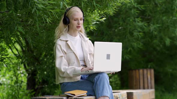 Young Woman in Big Black Headphones Talks Video Call Using Laptop on a Park Bench