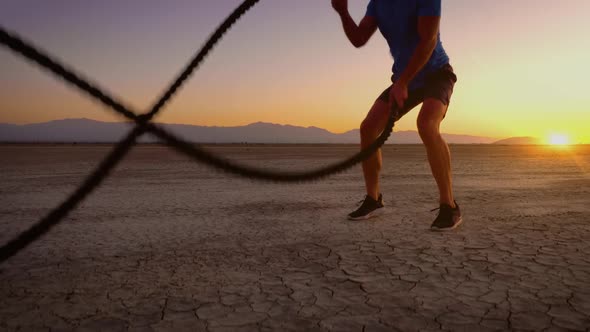 Athletic man working out with battle ropes on a dry lake at sunset alt