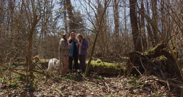 three girls stand forest holding a phone and a thermos in their hands, a white dog takes a stick alt