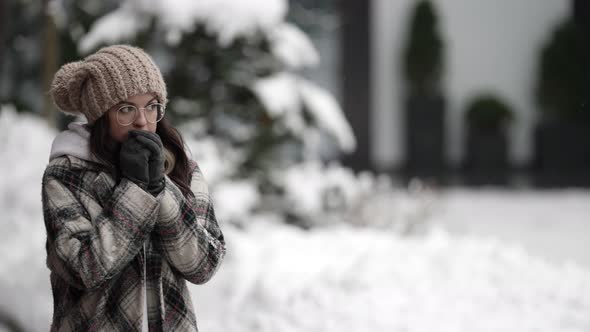 A Happy Young Darkhaired Woman in Round Glasses and a Plaid Coat Walks on a Snowy Winter Day Rubbing alt
