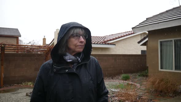 An elderly woman with glasses and a raincoat standing in a winter weather rain storm as raindrops fa alt