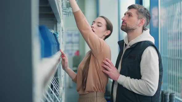 Side View of Charming Slim Beautiful Woman Choosing Pipe Connector with Man in Hardware Store alt