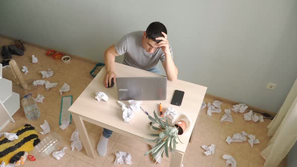 young man sits at table at home and recalculates coins . Around him, chaos and strong mess. alt