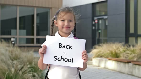 Cute Smiling Little Schoolgirl in Uniform Stands with a Poster Back To School Near the School alt