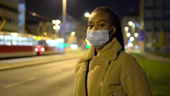 A Young Black Woman in a Face Mask Looks at the Camera As She Stands By a Road in a Street at Night alt