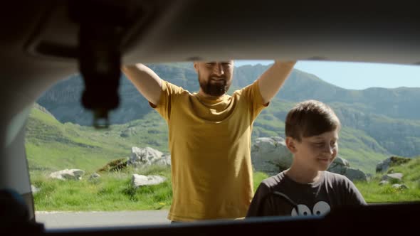 Happy father and son open car trunk and take backpack gets things out against mountain landscape alt