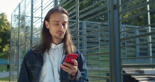 Crop View of Long Haired Guy Using Smartphone While Walking Near Sports Ground. Millennial Man with alt