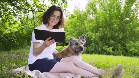 Girl in Glasses Reads Book Sitting Near Dog on Grass