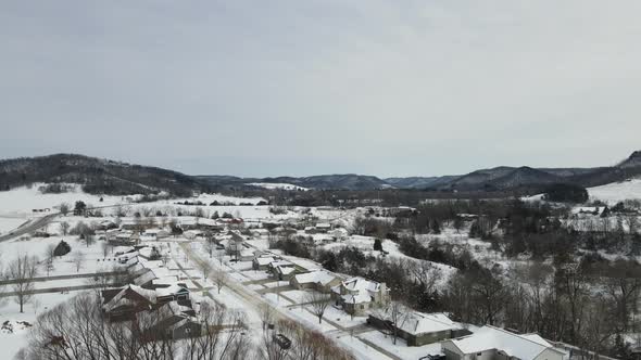 Flyover of rural neighborhood in valley covered in snow. Mountains and forests in the background.