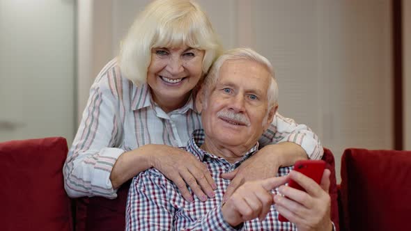 Happy Smiling Senior Couple with Mobile Phone at Home. Resting on Sofa in Cozy Living Room alt
