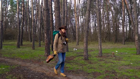 A Backpacker with a Guitar and a Camera Walks Along a Trail in the Forest alt