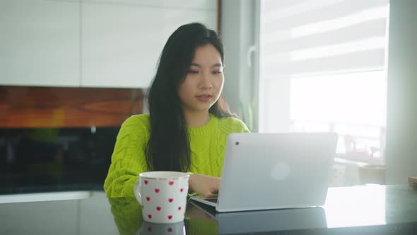 Young Asian Woman Working Remotely. Freelancer Typing on the Laptop with Cup of Tea or Coffee in Her alt
