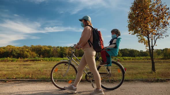 Mom with a Child in Medical Masks Walk with a Bicycle alt