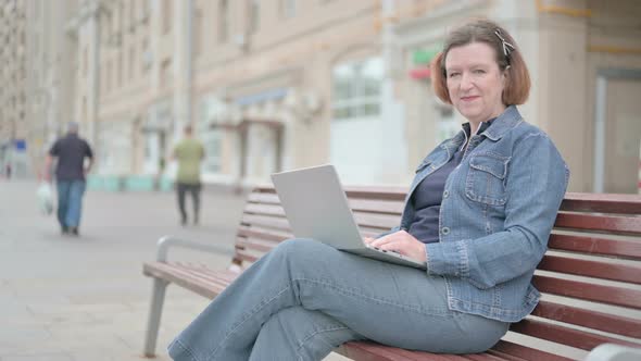 Old Woman with Laptop Showing Thumbs Up Sign While Sitting Outdoor on Bench alt
