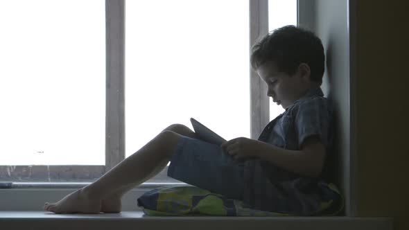 Cute Little Boy Uses A White Tablet Pc On A Windowsill At Home