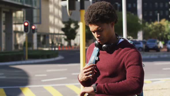 African american man in city checking smartwatch, wearing headphones and backpack waiting in street alt