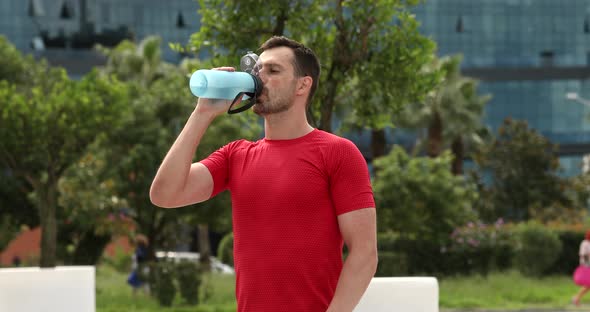 Young muscular man wearing red sports shirt drinking water from bottle.