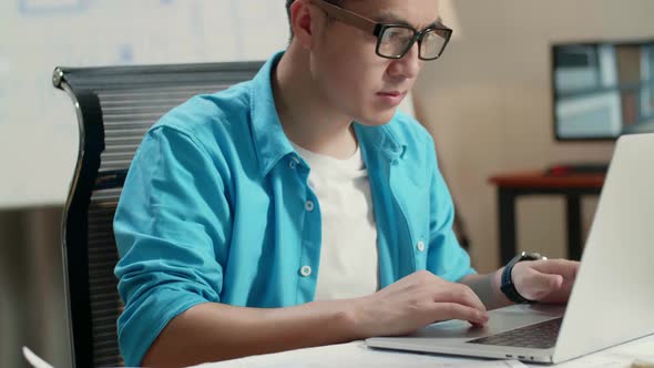 Close Up Of Asian Man Engineer Looking At Blueprint While Typing On A Laptop At The Office alt