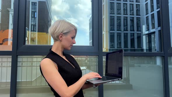 Beautiful Blonde Woman in a Black Dress is Typing on a Laptop in the City
