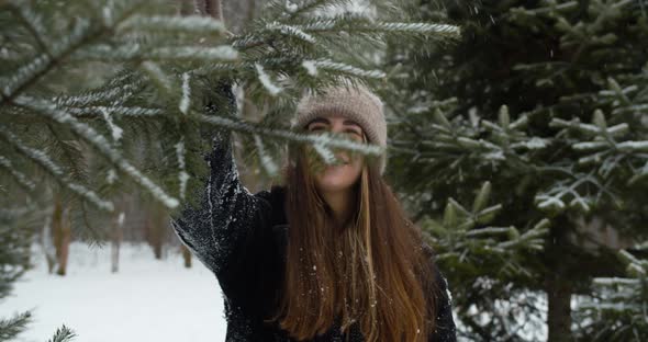 Playful Young Woman Shakes a Branch of a Christmas Tree with Snow and Smiles in Front of a Tree in