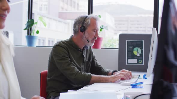 Caucasian businessman sitting using computer talking on phone headset in office alt