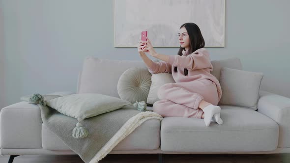 Young cheerful caucasian dark-haired woman with glasses sitting on sofa and taking selfie on phone.