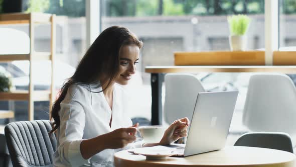 Pretty Worker in White Shirt Typing on Computer with Smile and Drinking From White Cup alt