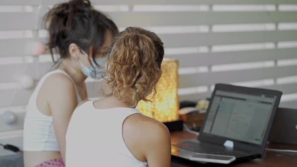 Two Young Women Wearing a Protective Mask Sitting at Table in Cafe with Laptop Works Together alt