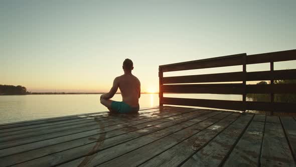 A Young Man is Sitting on the Edge of the Pier and Meditating alt
