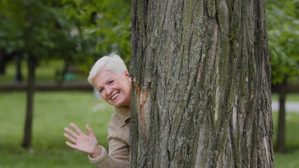Cheerful Mature Middle Aged Woman Peeks Out From Behind Large Tree Plays Hide and Seek Smiling Older alt