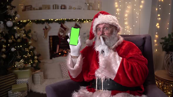 Happy Santa Claus Sitting in Chair Near Christmas Tree and Fireplace, Showing Mobile Phone