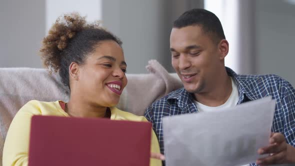 Positive Laughing African American Couple Analyzing Diagrams Talking Sitting on Couch in Home Office alt