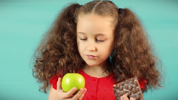 Child Makes a Choice Between of Holding a Green Apple and Chocolate Cute Child on Turquoise alt