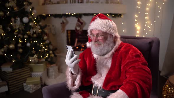 Santa Claus Is Sitting in a Chair in Front of the Fireplace at Night and Using the Phone. Christmas