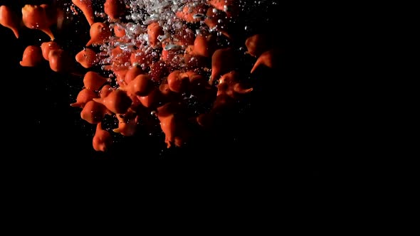 Beak peppers dropped into water on black background, side lit slow motion alt