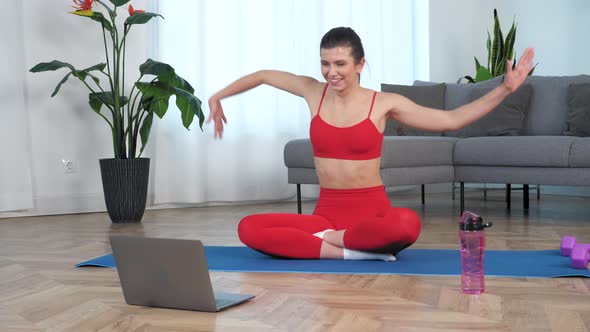 Smiling Woman in Sportswear Sitting on Yoga Mat Makes Wave with Hands at Home alt