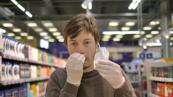 Man in a Medical Mask and Rubber Gloves in a Supermarket Removes the Mask and Scratches His Nose and alt