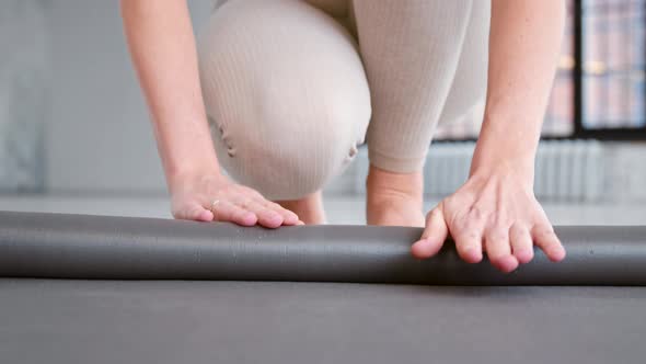 Young woman fitness trainer in light beige tracksuit rolls new rubber mat on floor alt