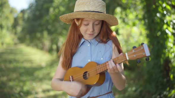 Medium Shot Portrait of Happy Skilled Caucasian Redhead Girl Singing Playing Ukulele Outdoors alt