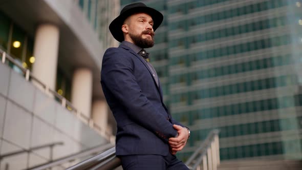 Stylish Mustache Man in Luxury Suit and Hat is Standing Alone in Modern Business District alt