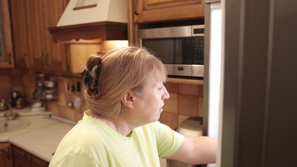Hungry middle aged woman opens a refrigerator at night and eating hamburger with sausage.